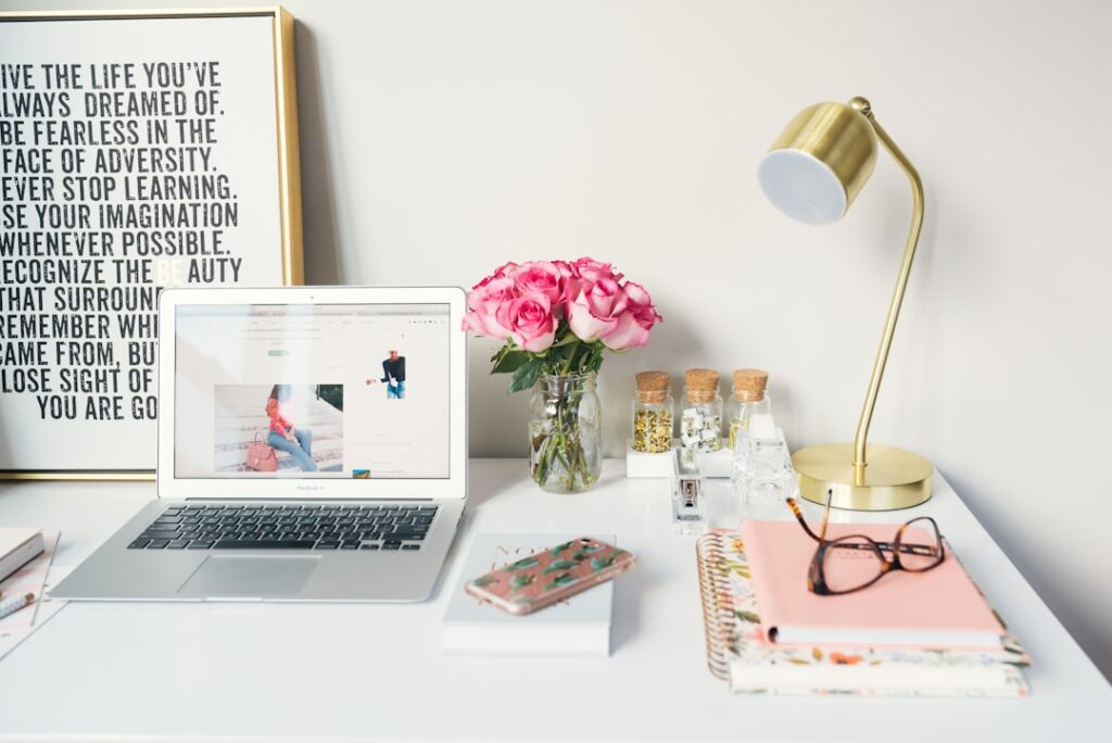 Ordinateur et bureau organisé avec des outils du quotidien Photo d'un bureau comprenant un ordinateur portable, un téléphone, une paire de lunettes, des fleurs et une lampe, symbolisant un espace de travail moderne et inspirant.